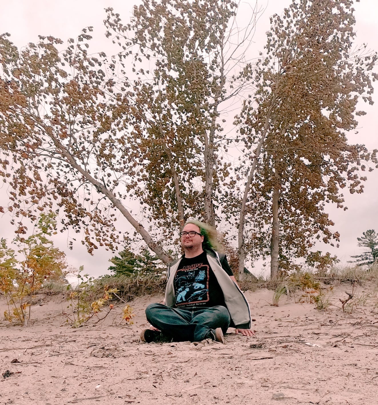 A man with long green hair sits on a beach in front of a short tree growing out of the sand